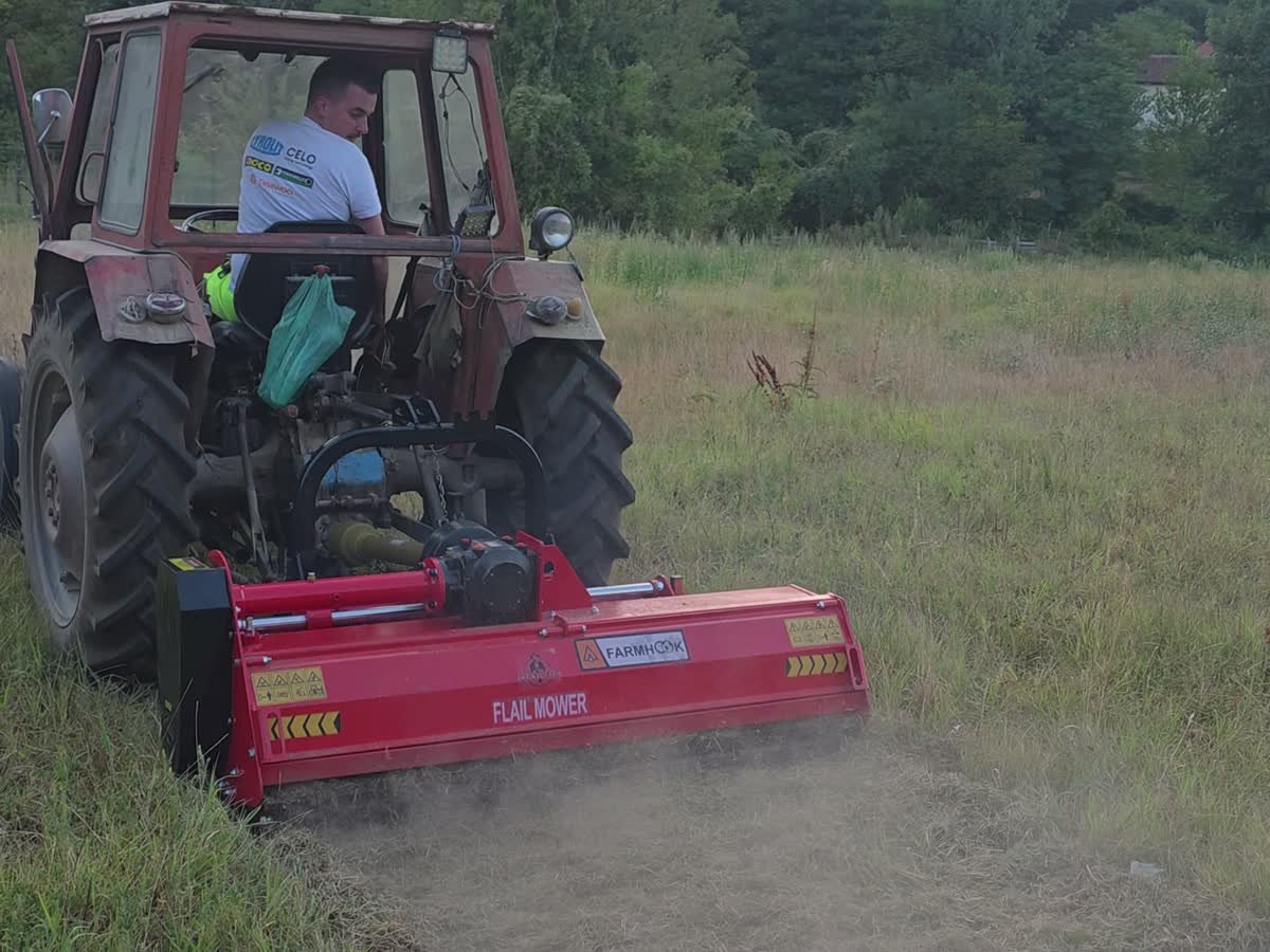 Traktor sa crvenim malčerom kosi visoku travu na livadi uz šumu