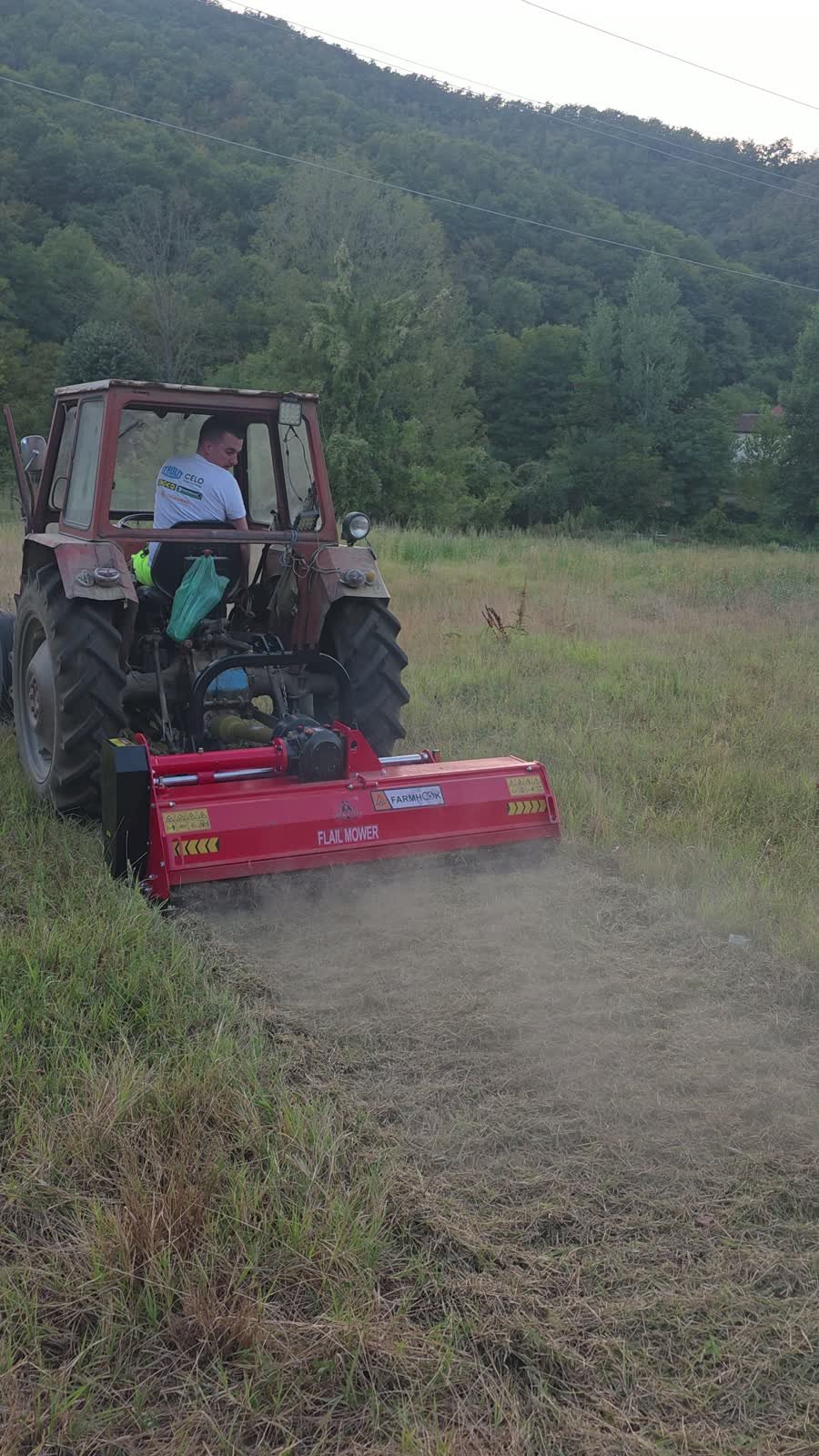 Traktor sa crvenim malčerom kosi visoku travu na livadi uz šumu